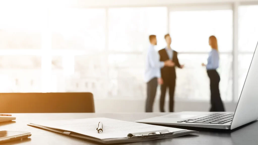Business professionals discussing in the background with a notebook and pen in the foreground.